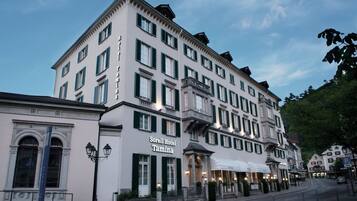 Een meerlaags hotel in klassieke Europese stijl, met een opvallende ingang met een luifel, meerdere ramen met luiken en een zichtbaar hotelbord.