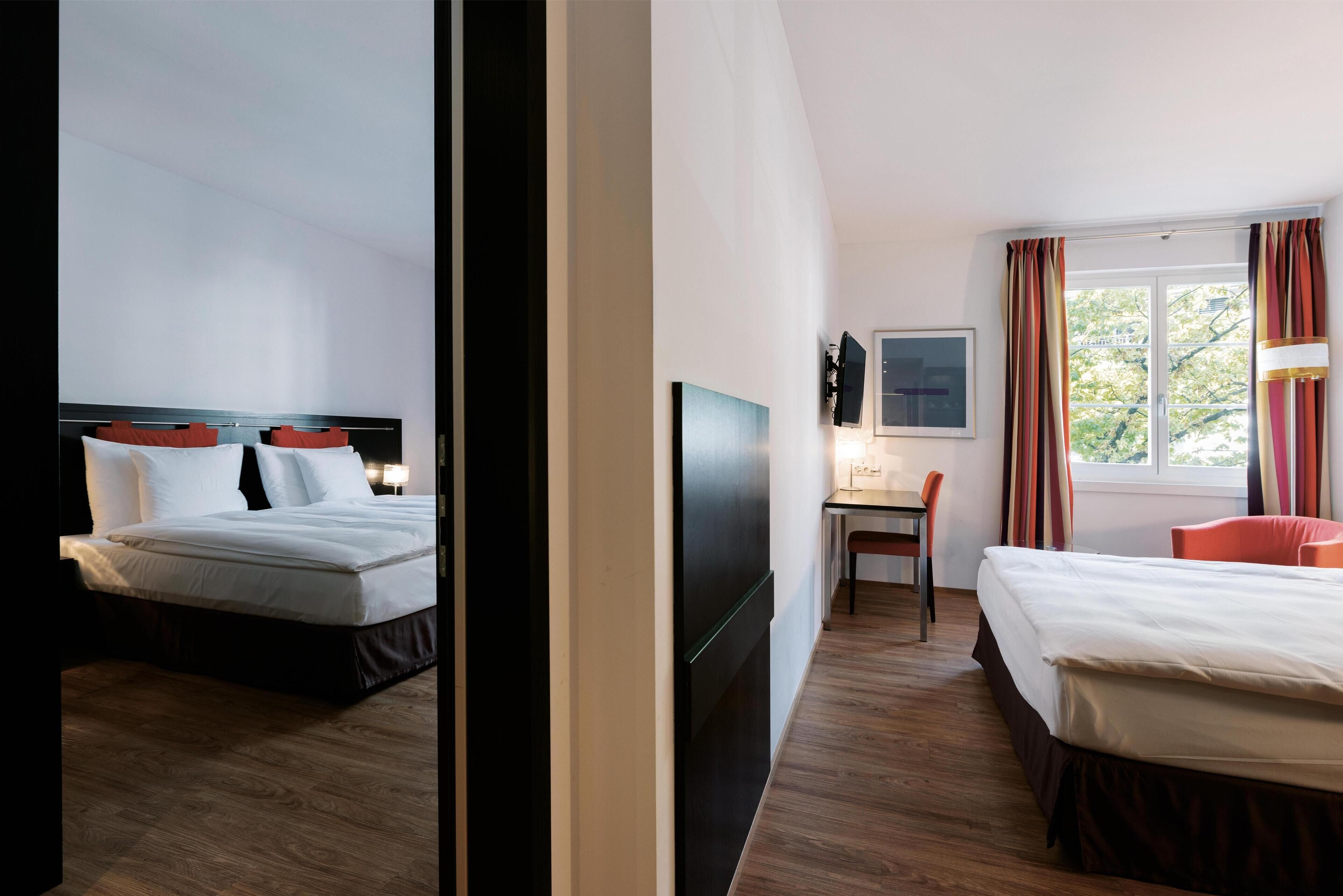 A hotel room with two beds, a desk, a chair, a TV, and a window with curtains.