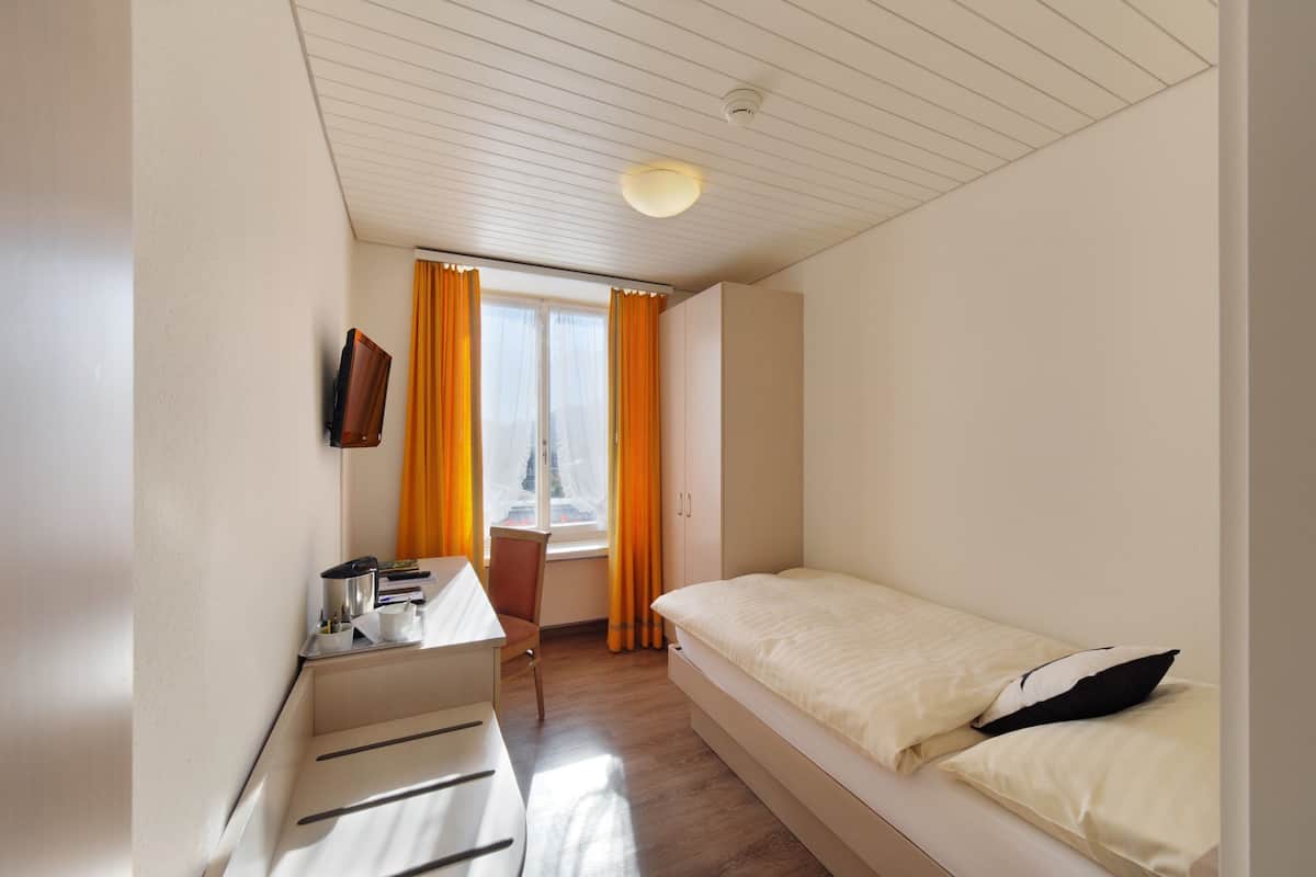 A small, well-lit bedroom with a single bed, a desk, and a chair.