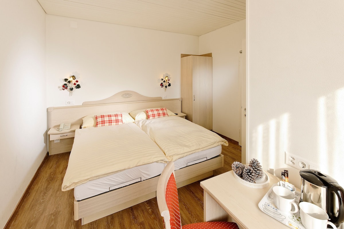 A double bed with a wooden headboard, a bedside table with a lamp, and a small table with cups and a kettle.