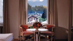 Ein Hotelzimmer mit Blick auf einen Berg und ein Dorf. Es gibt einen runden Tisch mit zwei Stühlen und eine Blumenvase.