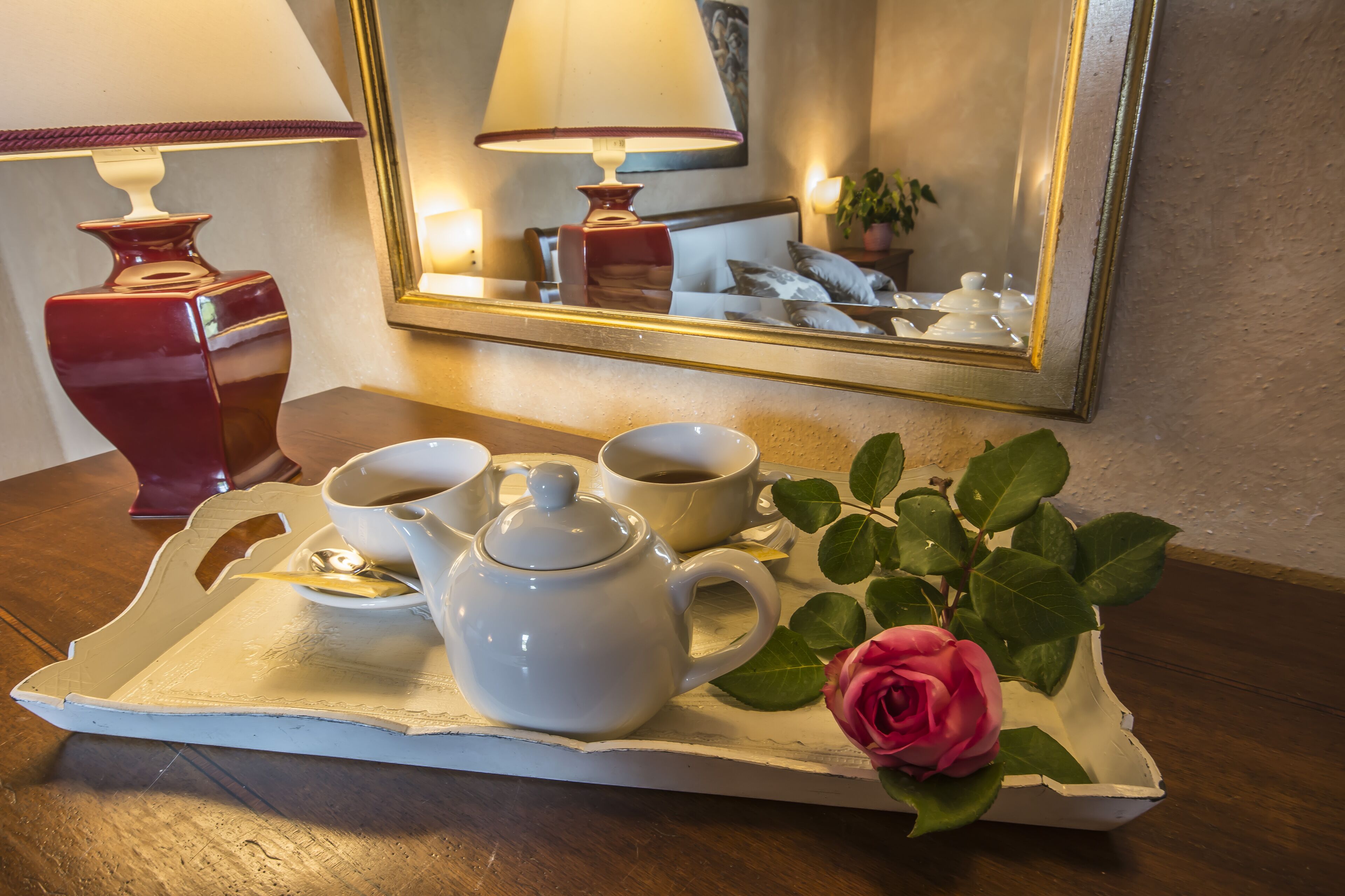 A tray with a teapot, cups, and saucers, a rose, and a mirror reflecting a lamp and a bed.