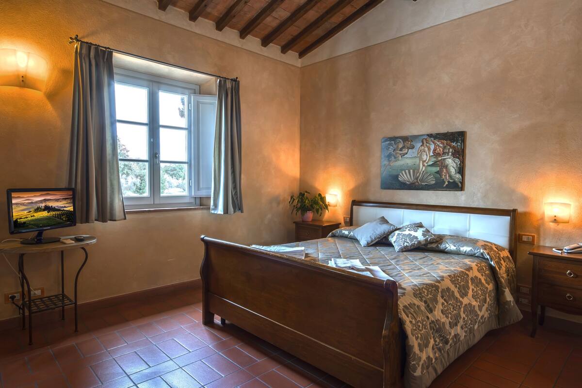 Un dormitorio con cama de madera, una ventana con cortinas, una mesita de noche con lámpara y un cuadro en la pared.