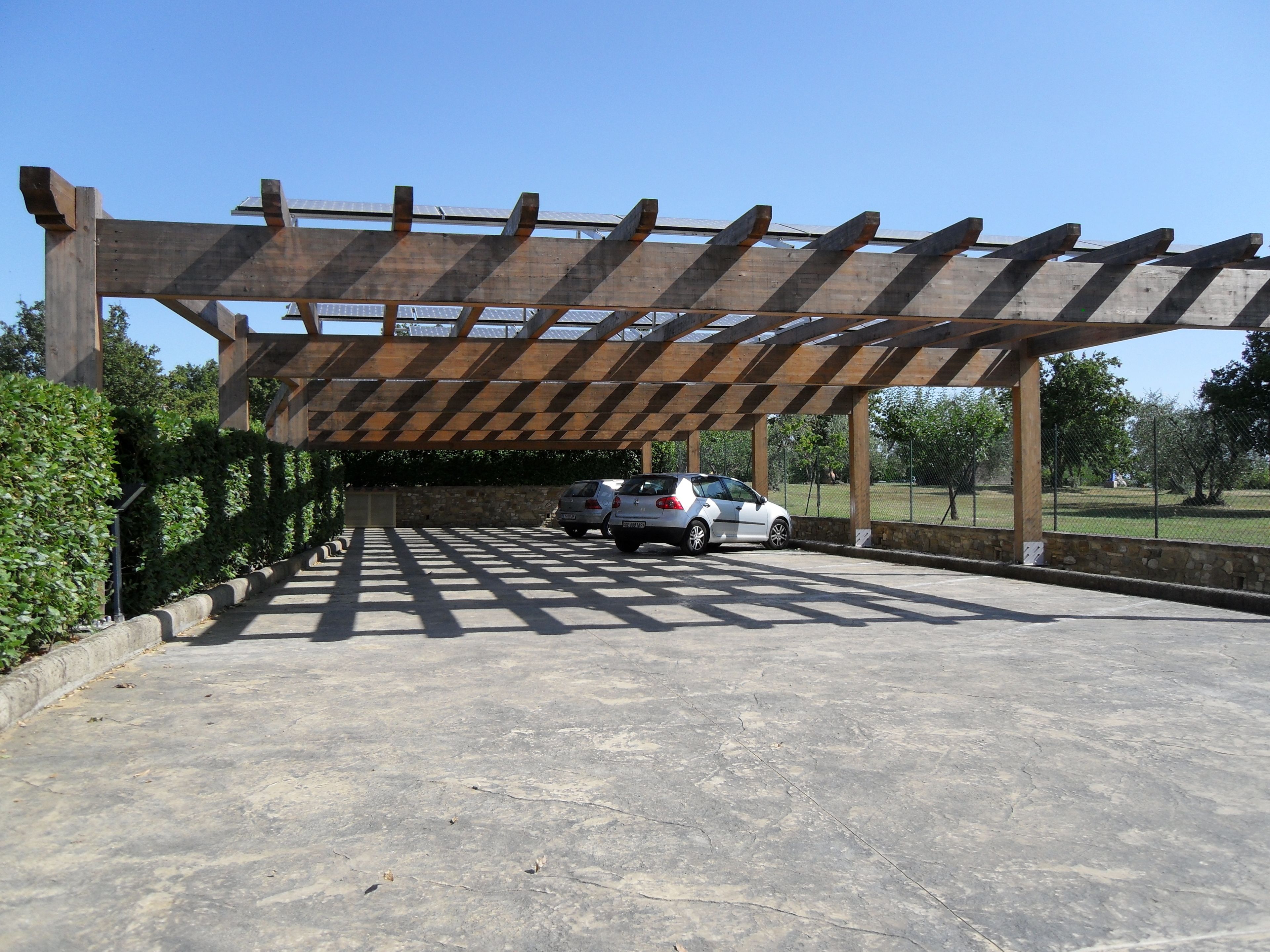 A covered parking area with a pergola structure, two cars parked, and a stone wall.
