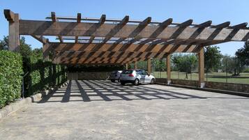 A covered parking area with a pergola structure, two cars parked, and a stone wall.