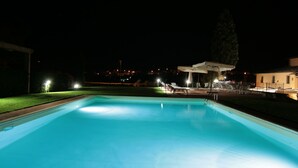 A nighttime swimming pool with a clear view of the surrounding area, including a covered seating area and distant buildings.