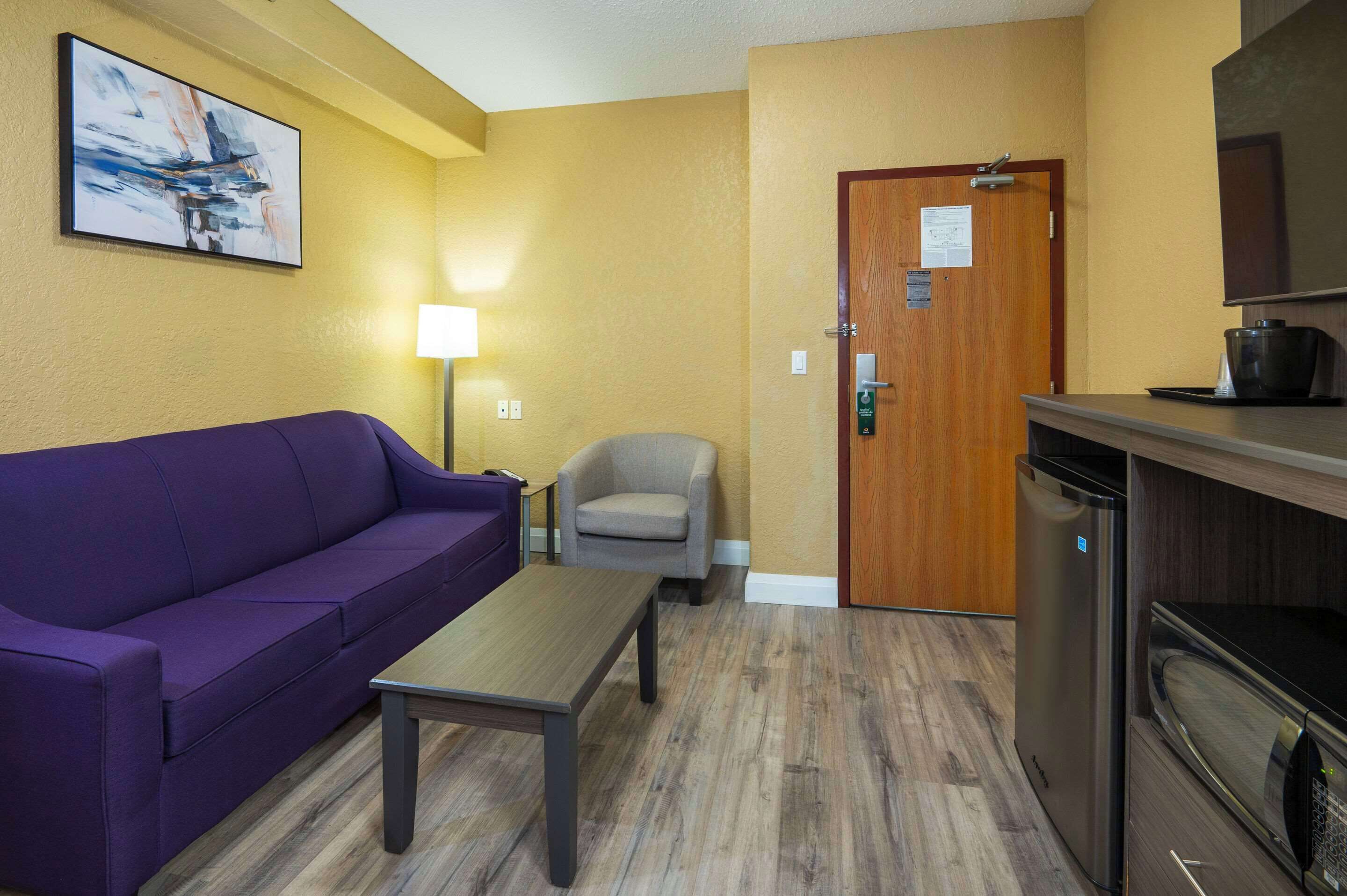 A room with a purple sofa, a small table, a chair, a wooden door, and a kitchenette with a microwave.