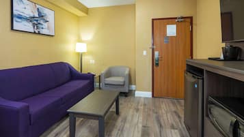 A room with a purple sofa, a small table, a chair, a wooden door, and a kitchenette with a microwave.