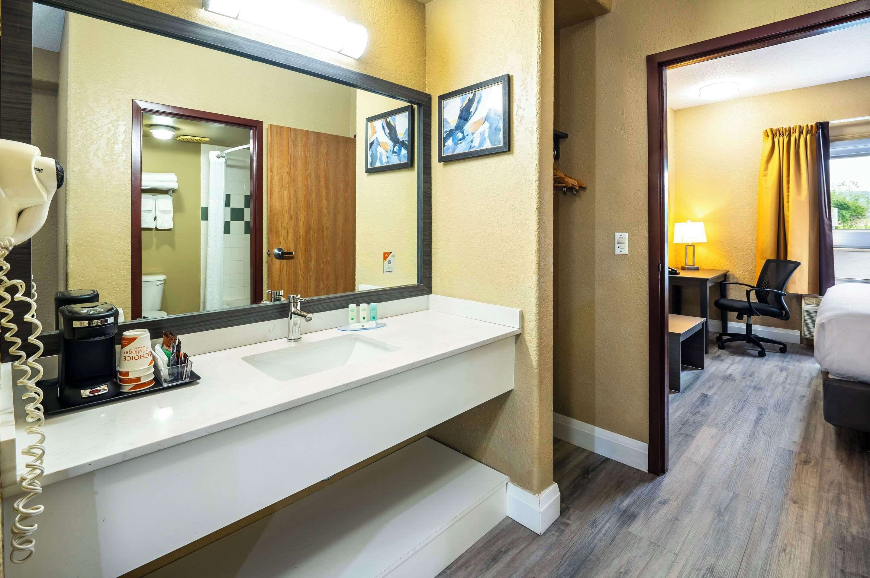 A hotel bathroom with a large mirror, a sink, and a hairdryer. There is a small table with toiletries and a coffee maker. The bathroom has wooden flooring and beige walls.