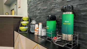 En hotellobby med en vägg av svart sten, en receptionsdisk och en kaffestation med en grön vattenautomat märkt med texten ”Varmt vatten” och en kaffemaskin.