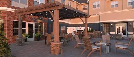 An outdoor patio area with wooden chairs and tables, a pergola with string lights, and a brick building in the background.
