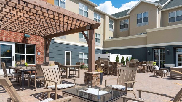 An outdoor patio area with wooden furniture, a glass-top table, and a stone planter. - Homewood Suites by Hilton Indianapolis-Airport/Plainfield (Plainfield)