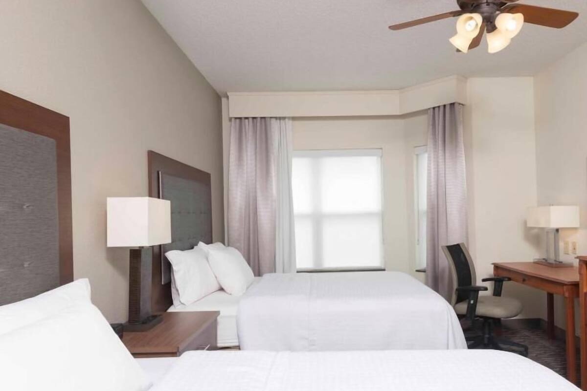 A hotel room with two beds, a desk, a chair, a ceiling fan, and a window with curtains.