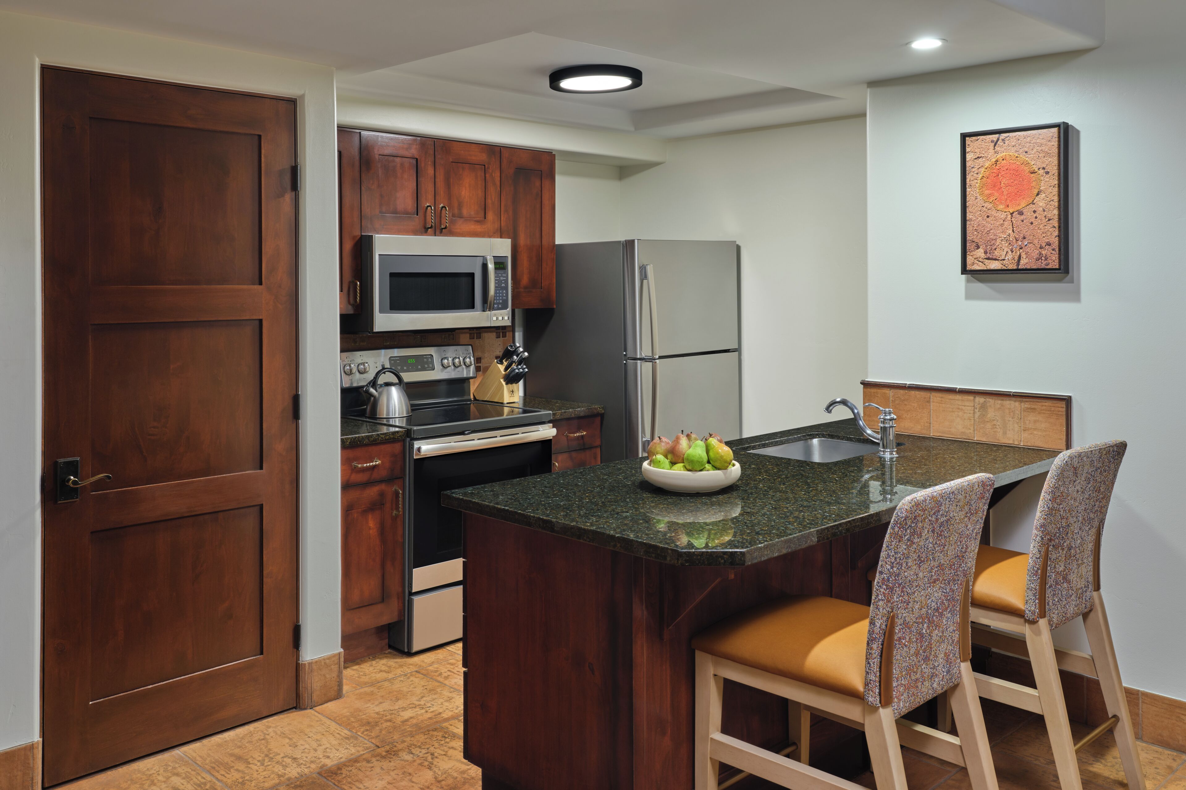 A kitchen with wooden cabinets, a stainless steel refrigerator, a microwave, and a sink. There is a fruit bowl on the counter and two bar stools.