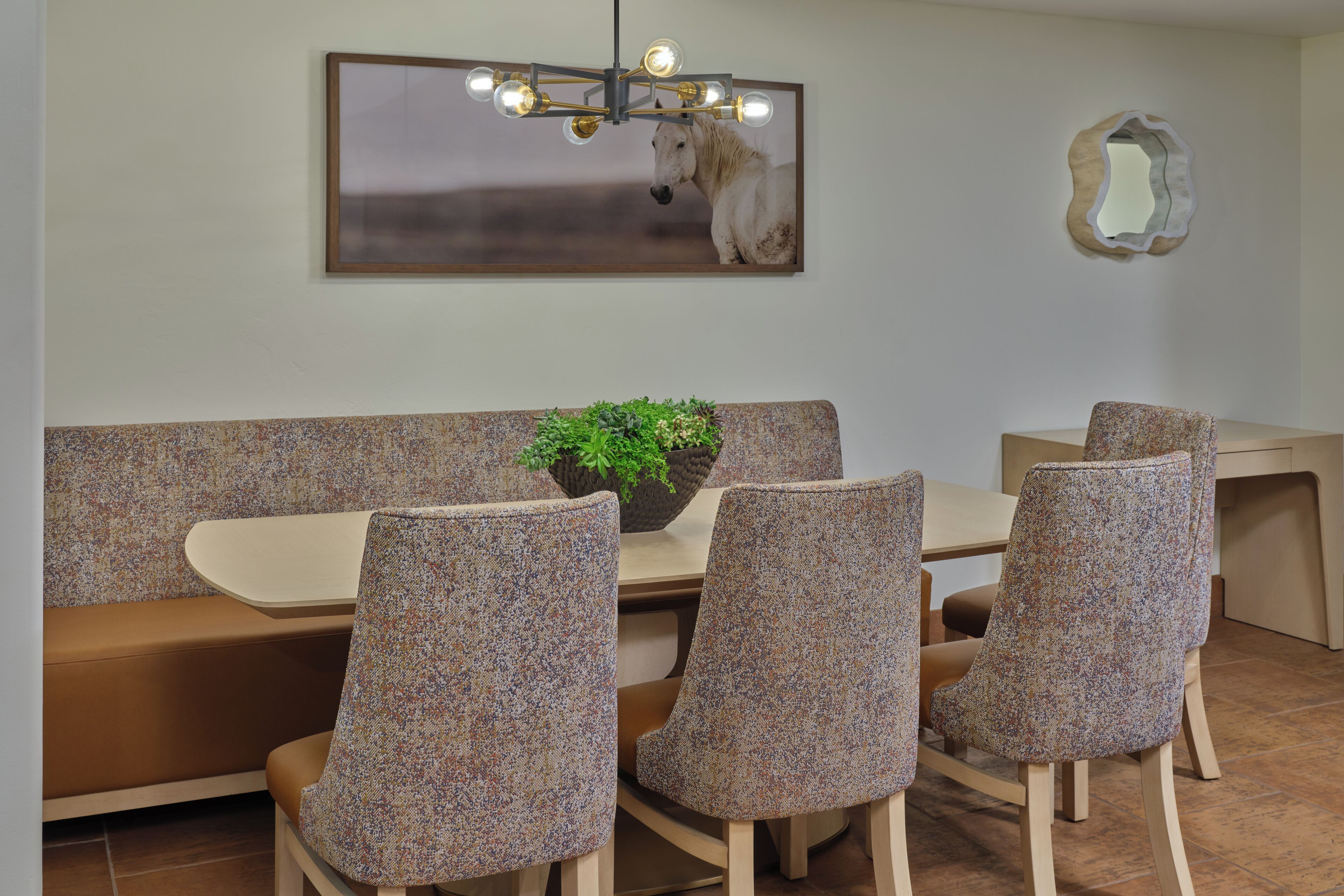 Un comedor con un banco, cuatro sillas tapizadas, una mesa, una planta en maceta, un espejo fijado a la pared y un cuadro con la imagen de un caballo.