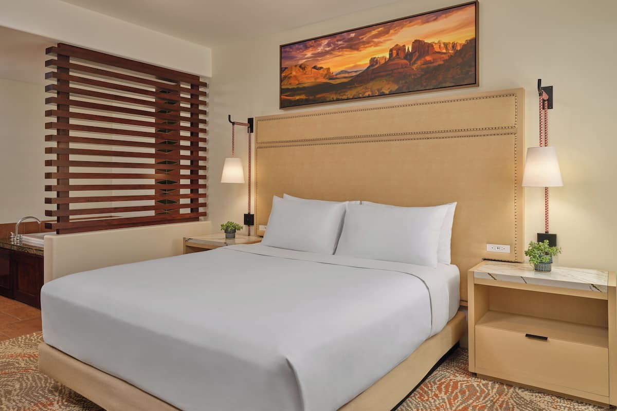 Een hotelkamer met een bed, nachtkastjes, een lamp, een schilderij en een raam met jaloezieën.