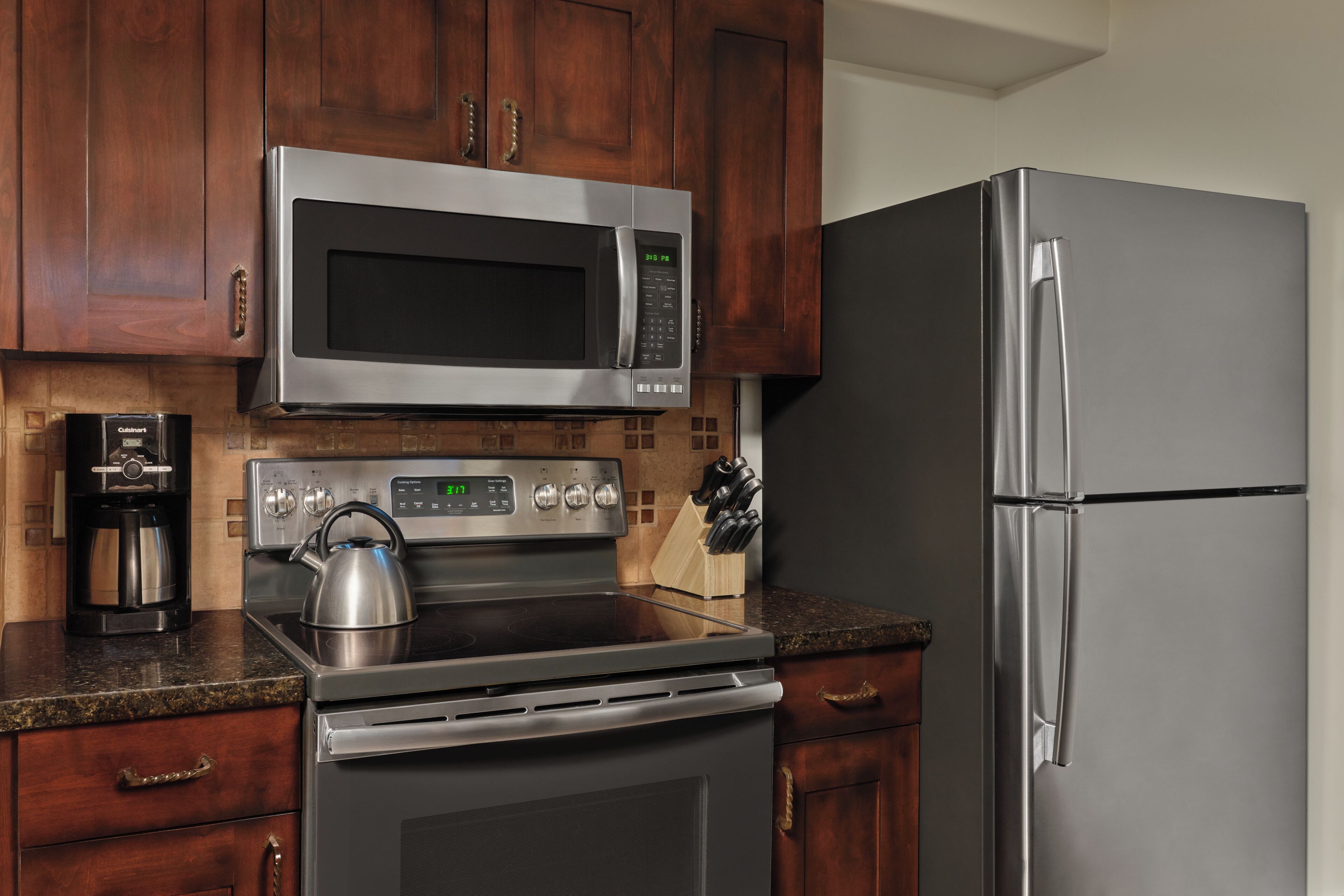A kitchen with a stainless steel microwave, oven, and refrigerator, wooden cabinets, and a granite countertop.