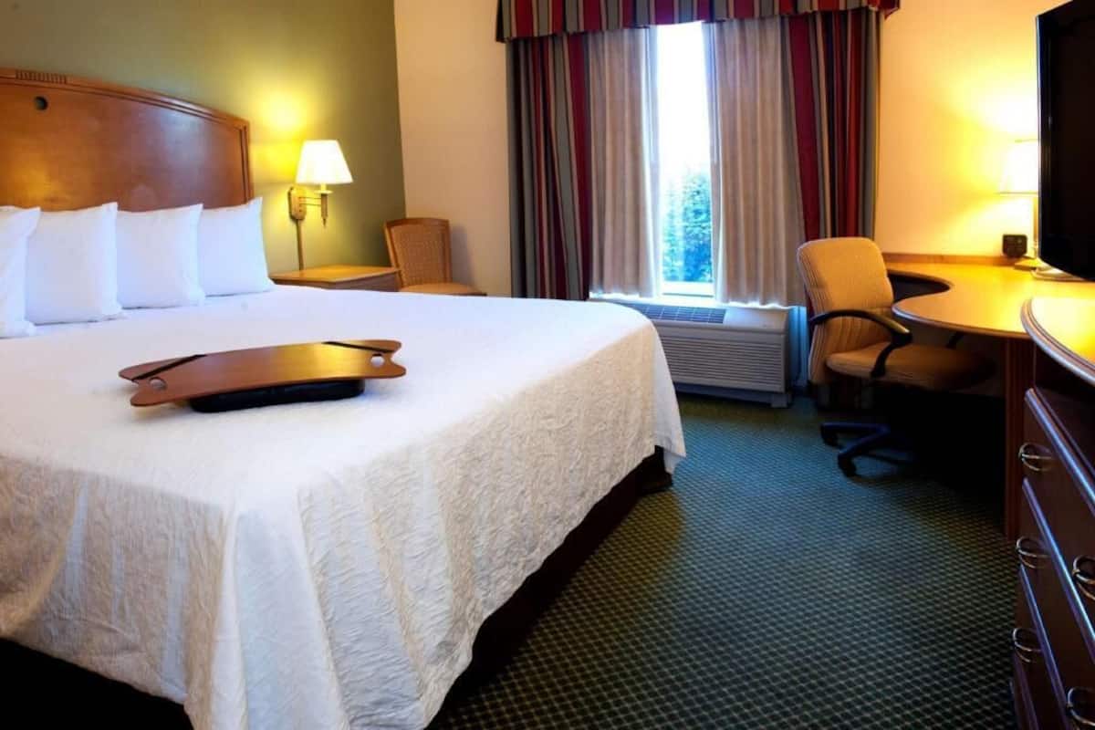 Quarto de hotel com uma cama grande, uma escrivaninha, uma cadeira, televisão e janela com cortinas.