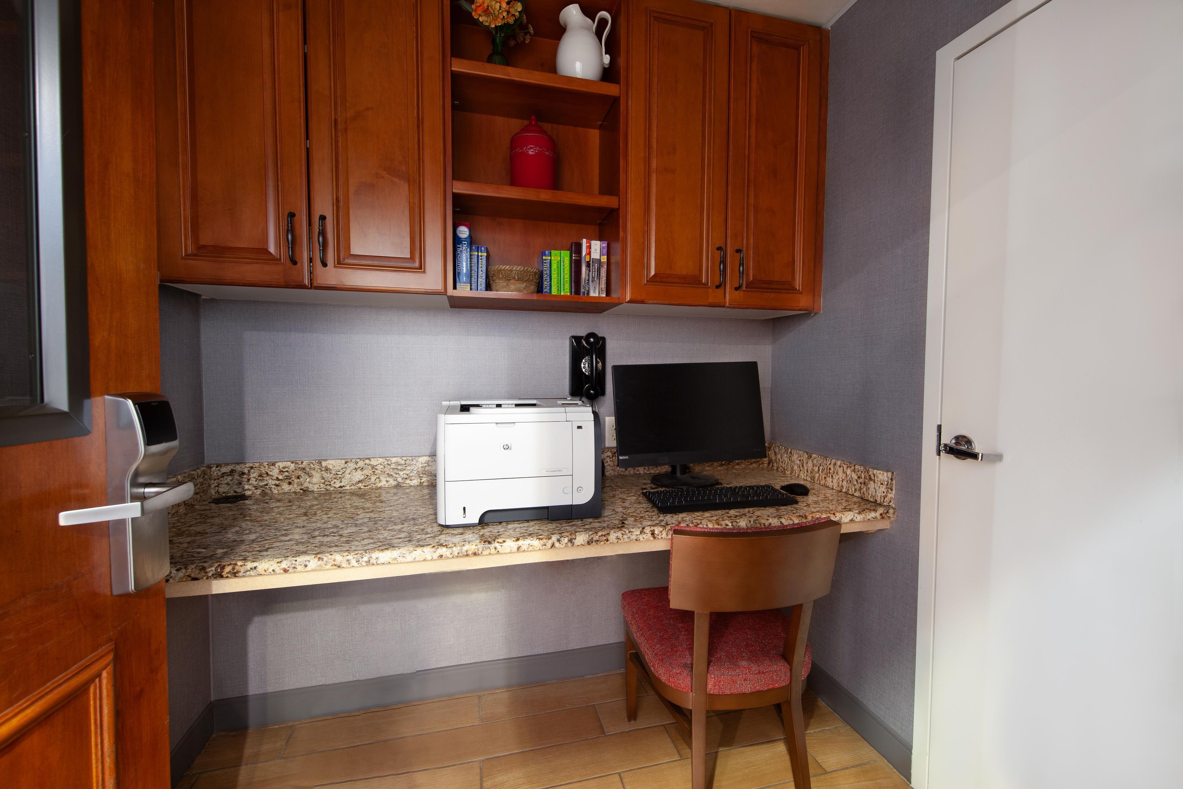Een kleine keuken met houten kastjes, een aanrecht, een printer, een computer monitor en een stoel.