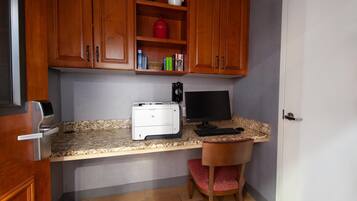 A compact kitchen with wooden cabinets, a countertop, a printer, a computer monitor, and a chair.