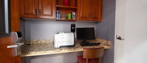 A compact kitchen with wooden cabinets, a countertop, a printer, a computer monitor, and a chair.