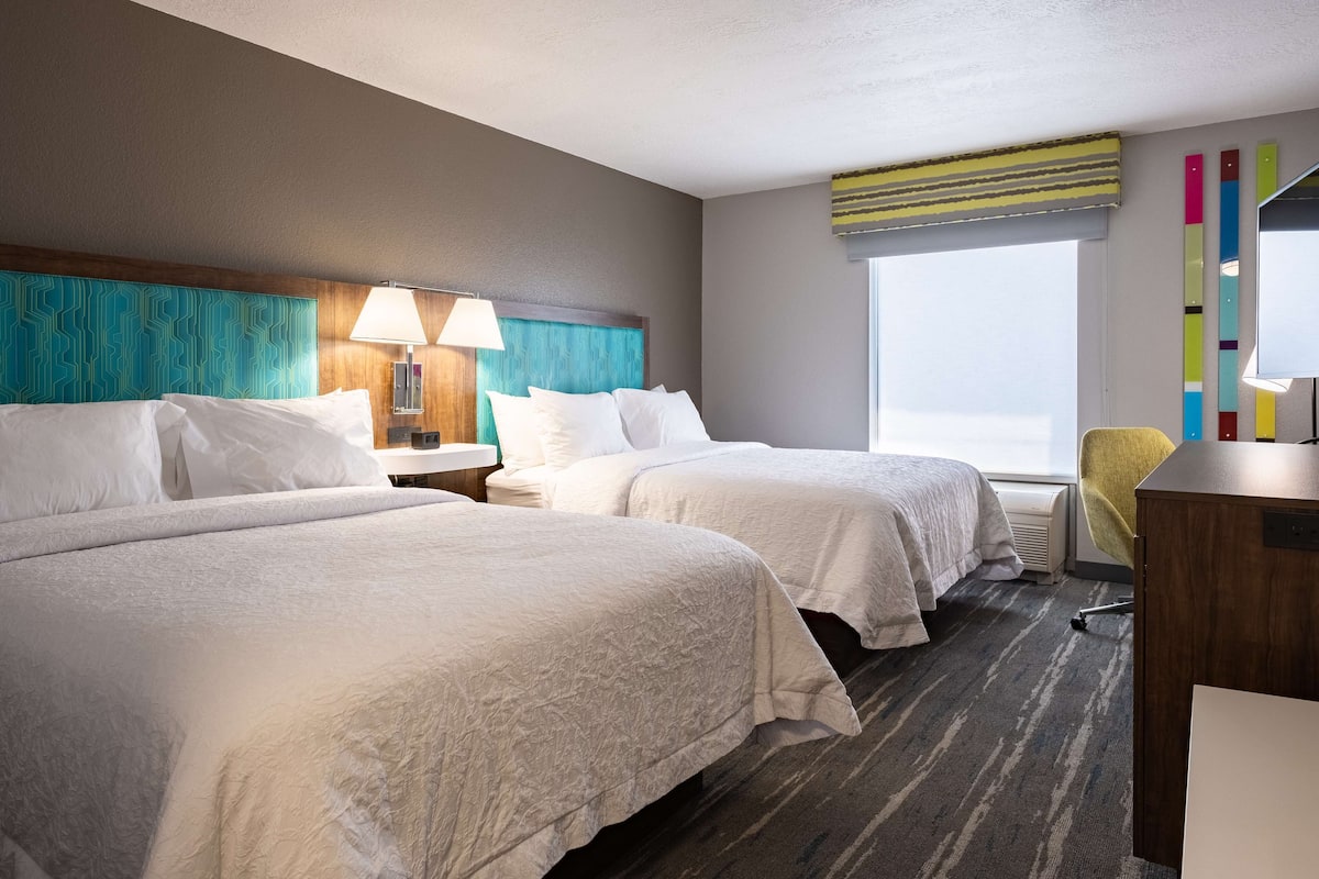 Quarto de hotel com duas camas, uma escrivaninha, uma cadeira e uma janela grande.