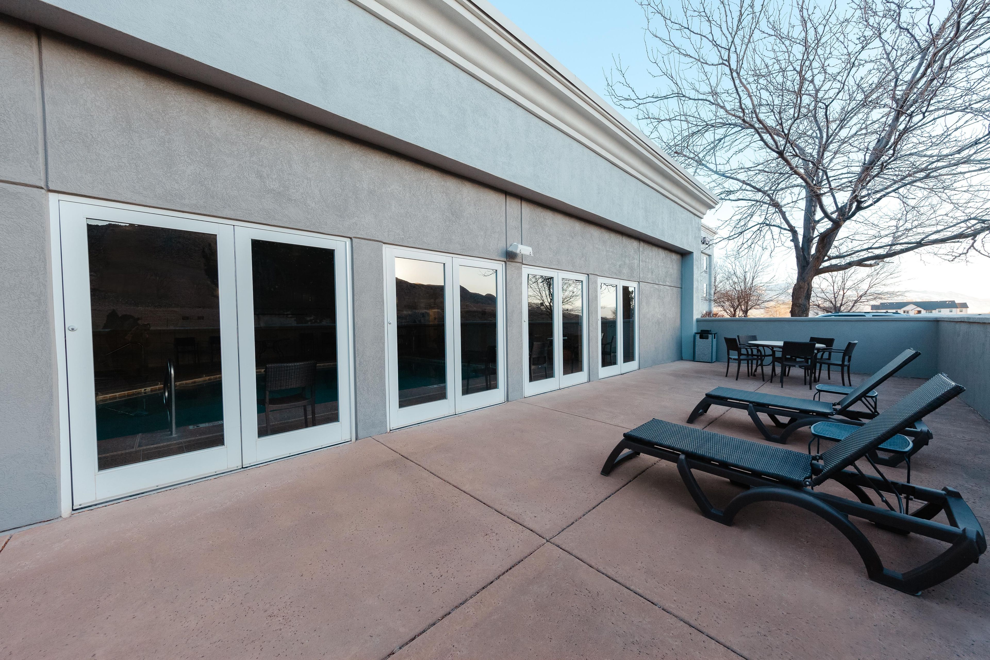A building with large sliding glass doors, outdoor seating with lounge chairs, and a tree in the background.