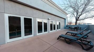 A building with large sliding glass doors, outdoor seating with lounge chairs, and a tree in the background.