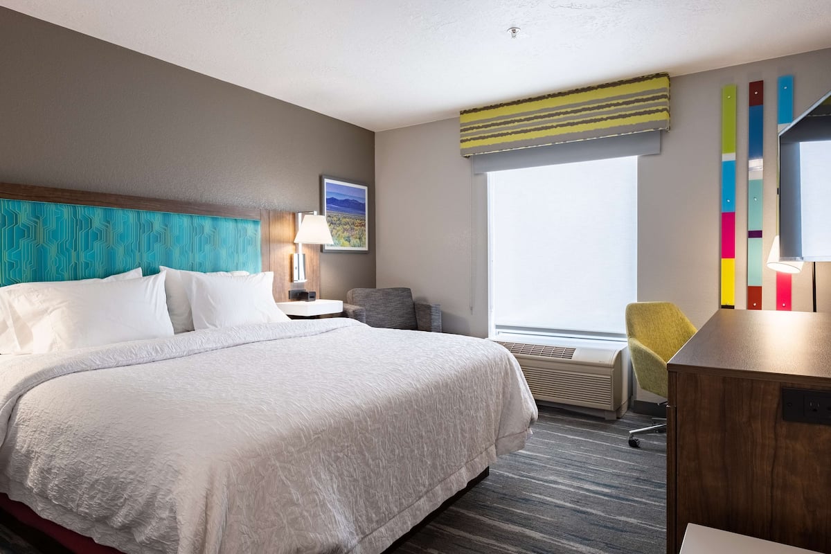 Quarto de hotel com uma cama grande, uma mesa, uma cadeira, janela com vista e um quadro na parede.