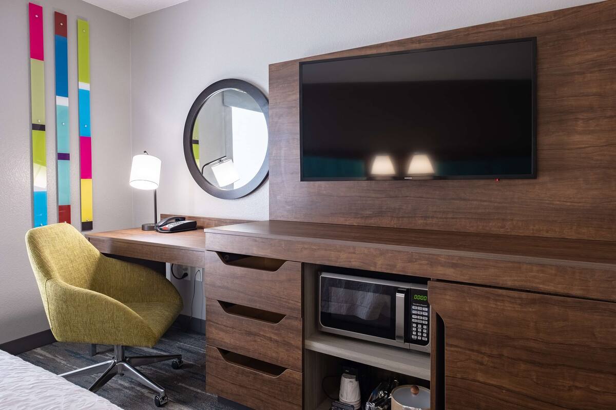 Quarto de hotel com escrivaninha de madeira, uma cadeira amarela, televisão de tela plana, espelho redondo, luminária e micro-ondas.