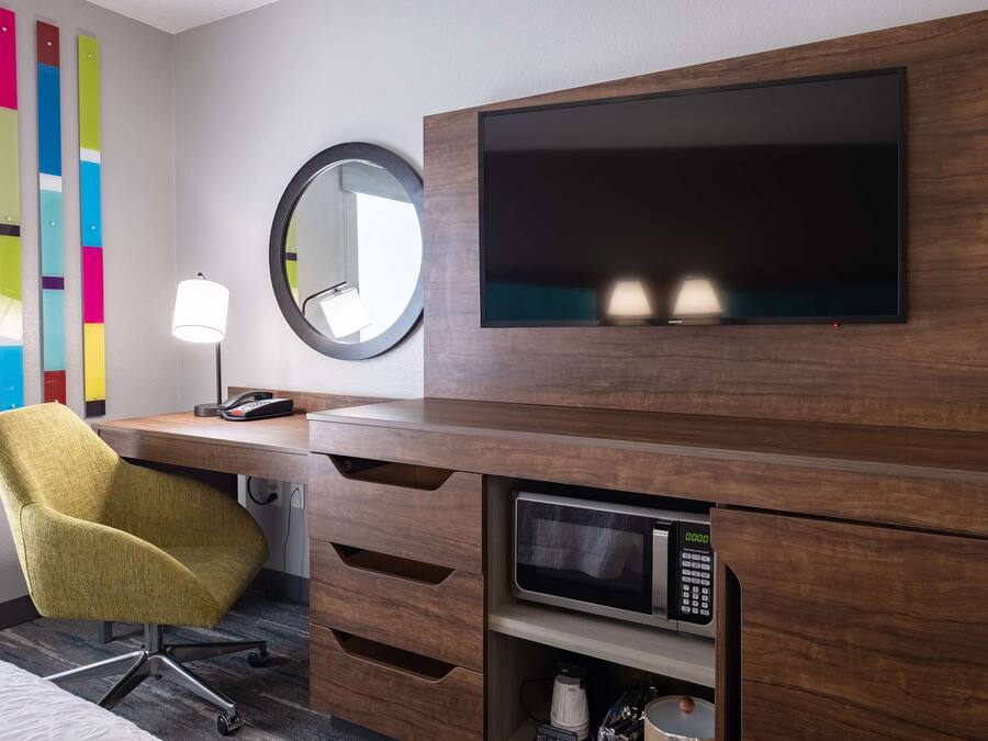 A hotel room with a wooden desk, a yellow chair, a flat-screen TV, a round mirror, a lamp, and a microwave.