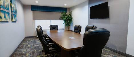 A conference room with a long table, six chairs, a potted plant, a flat-screen TV, and artwork on the walls.