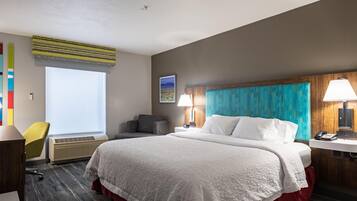 A hotel room with a large bed, a desk with a lamp, a chair, and a window with blinds.
