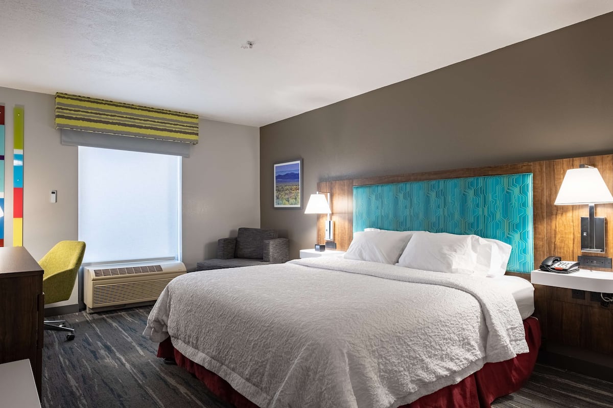Quarto de hotel com uma cama grande, uma escrivaninha, uma cadeira, uma janela com persianas, uma luminária na parede e um quadro.