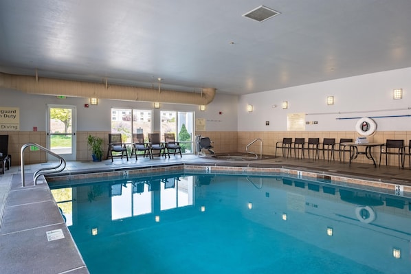 Una piscina interior con agua cristalina, rodeada de sillones de descanso y mesas, y un cartel de elevador para piscina.