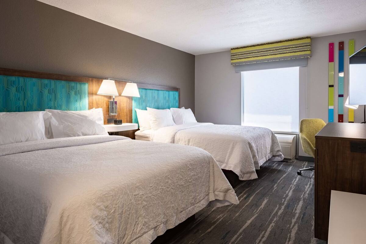 Quarto de hotel com duas camas, uma escrivaninha, uma cadeira, uma janela com vista e uma parede com padrão geométrico.