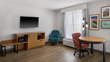 A hotel room with a flat-screen TV, a wooden cabinet with a microwave, a teal armchair, a red office chair, and a round table.
