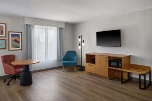 Una habitación de hotel moderna con piso de madera, una mesa redonda, una silla de escritorio roja, un sillón azul aguamarina, un armario de madera con microondas y un televisor de pantalla plana montado en la pared.