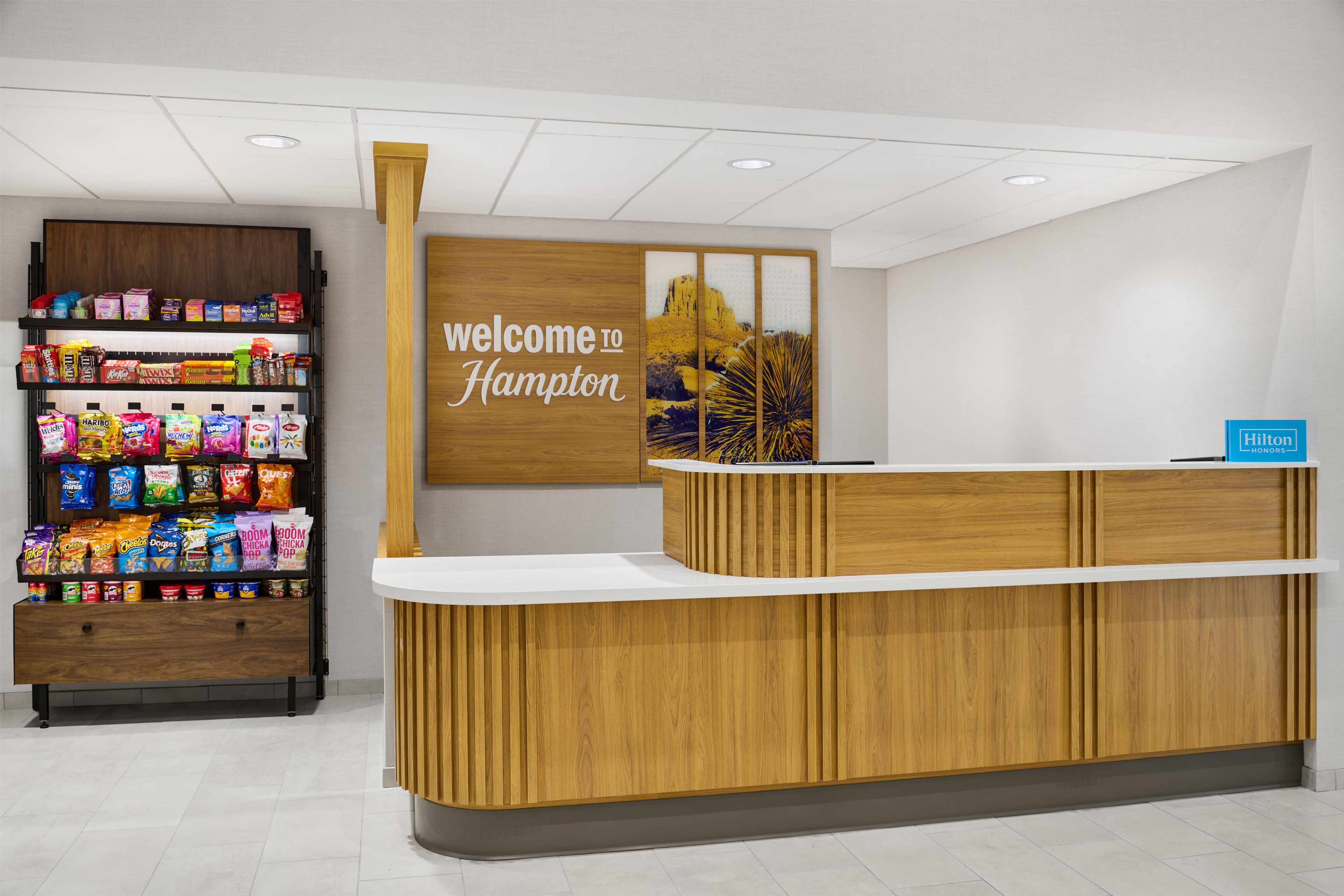 Uma mesa de recepção com a placa 'welcome to Hampton', prateleiras com lanches e a placa Hilton Honors.