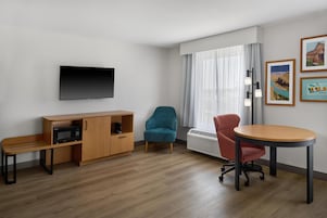 Habitación de hotel con televisión de pantalla plana, un mueble de madera con microondas, un sillón azul verdoso, una silla de escritorio roja y una mesa redonda.