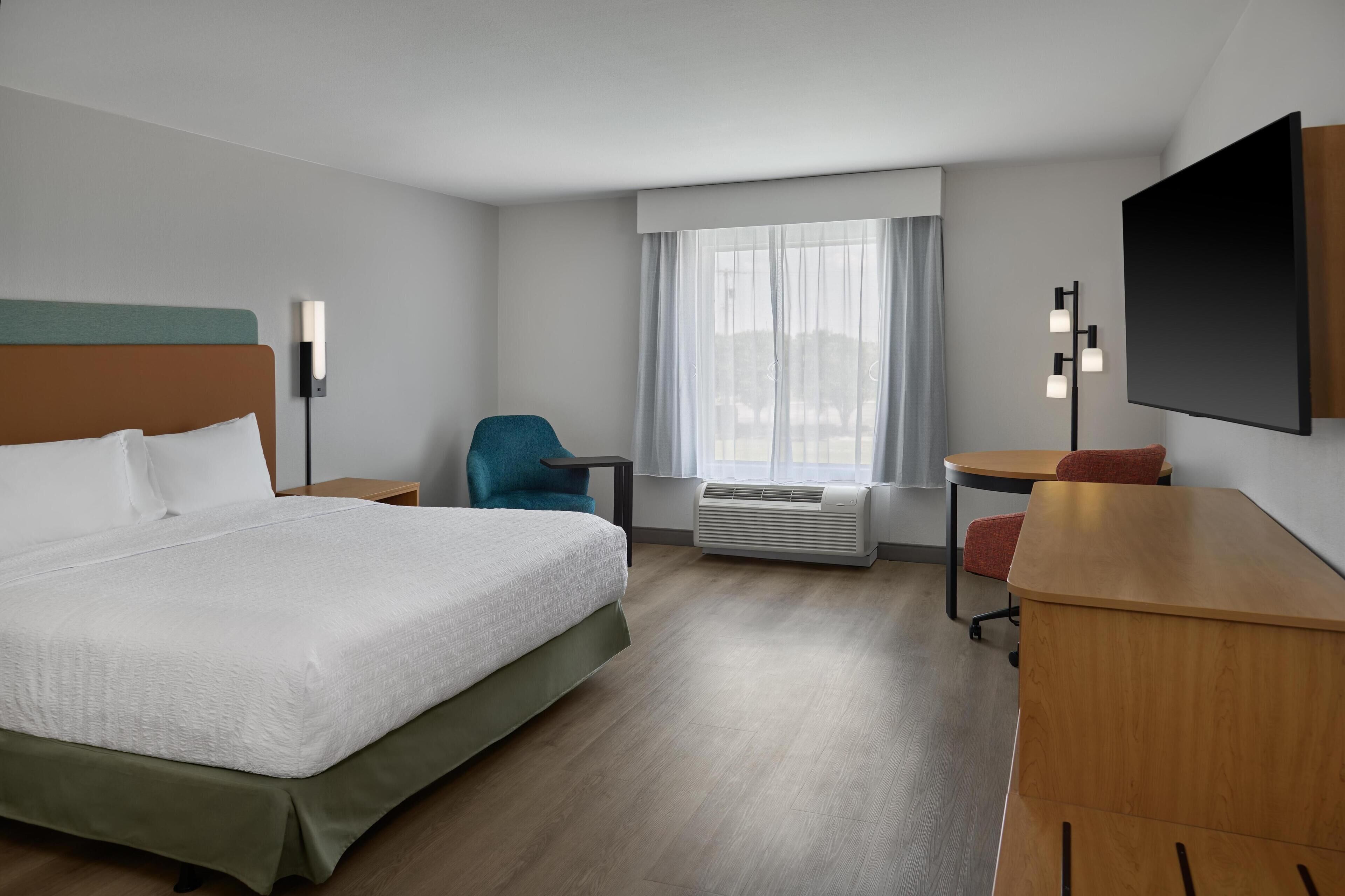 A hotel room with a bed, a desk, a chair, a TV, and a window with curtains.