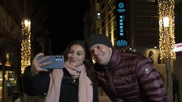 Duas pessoas tirando uma selfie em ambiente urbano, com edifícios iluminados e uma placa visível com a inscrição