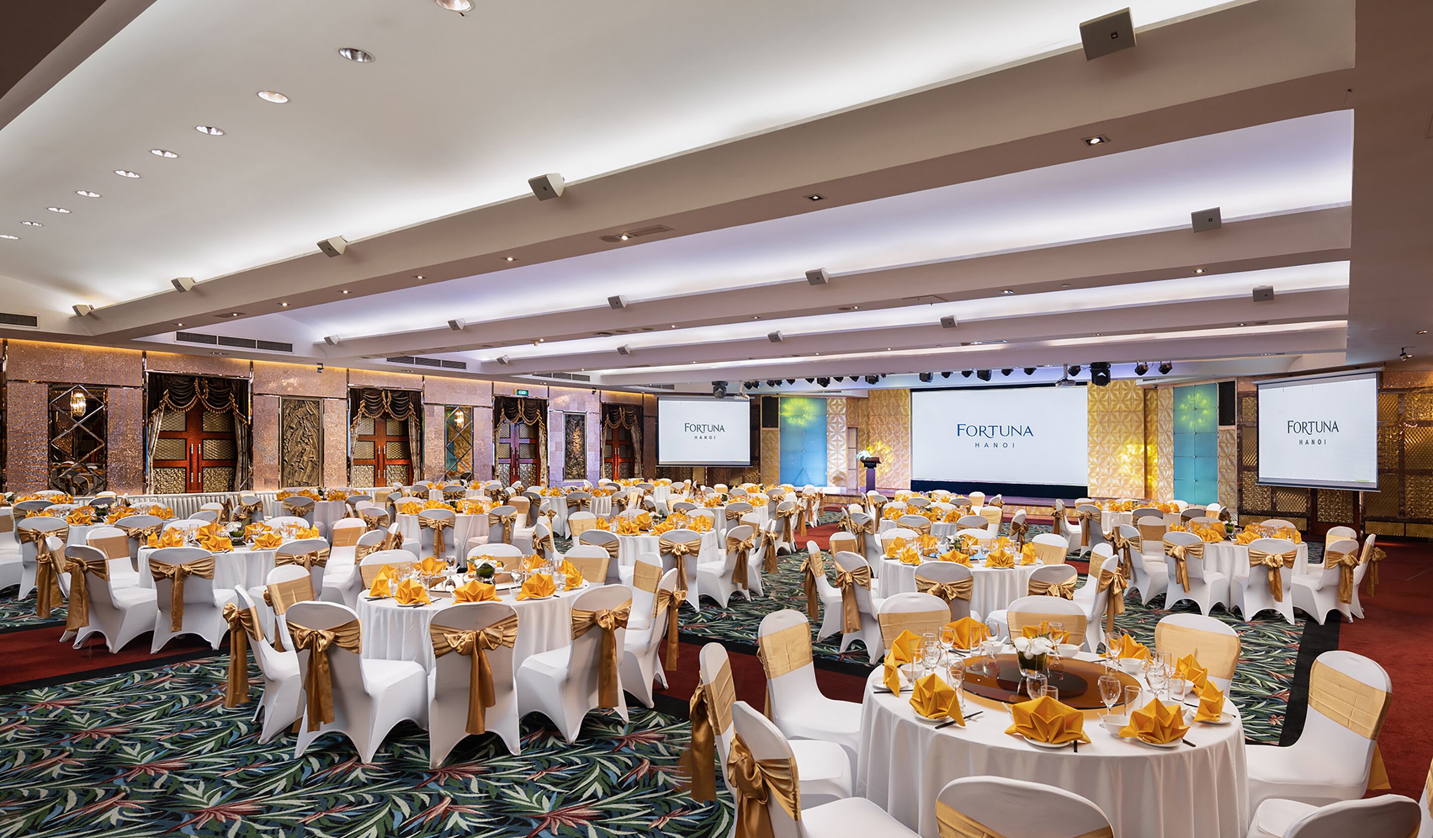 Un amplio salón de banquetes con filas de mesas redondas, cada una con mantel blanco y fajas doradas, y un escenario con una pantalla que muestra 'FORTUNA HANOI'.
