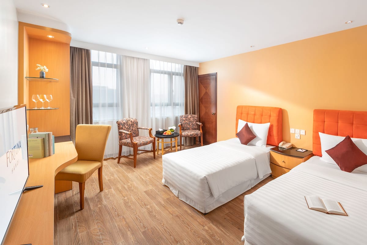 A hotel room with two beds, a desk, a chair, a small table with fruit, and a window with curtains.