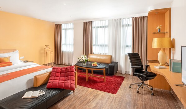 Habitación de hotel con cama, sofá rojo, sillón de cuero negro, una pequeña mesa con un jarrón de flores y un escritorio con lámpara.