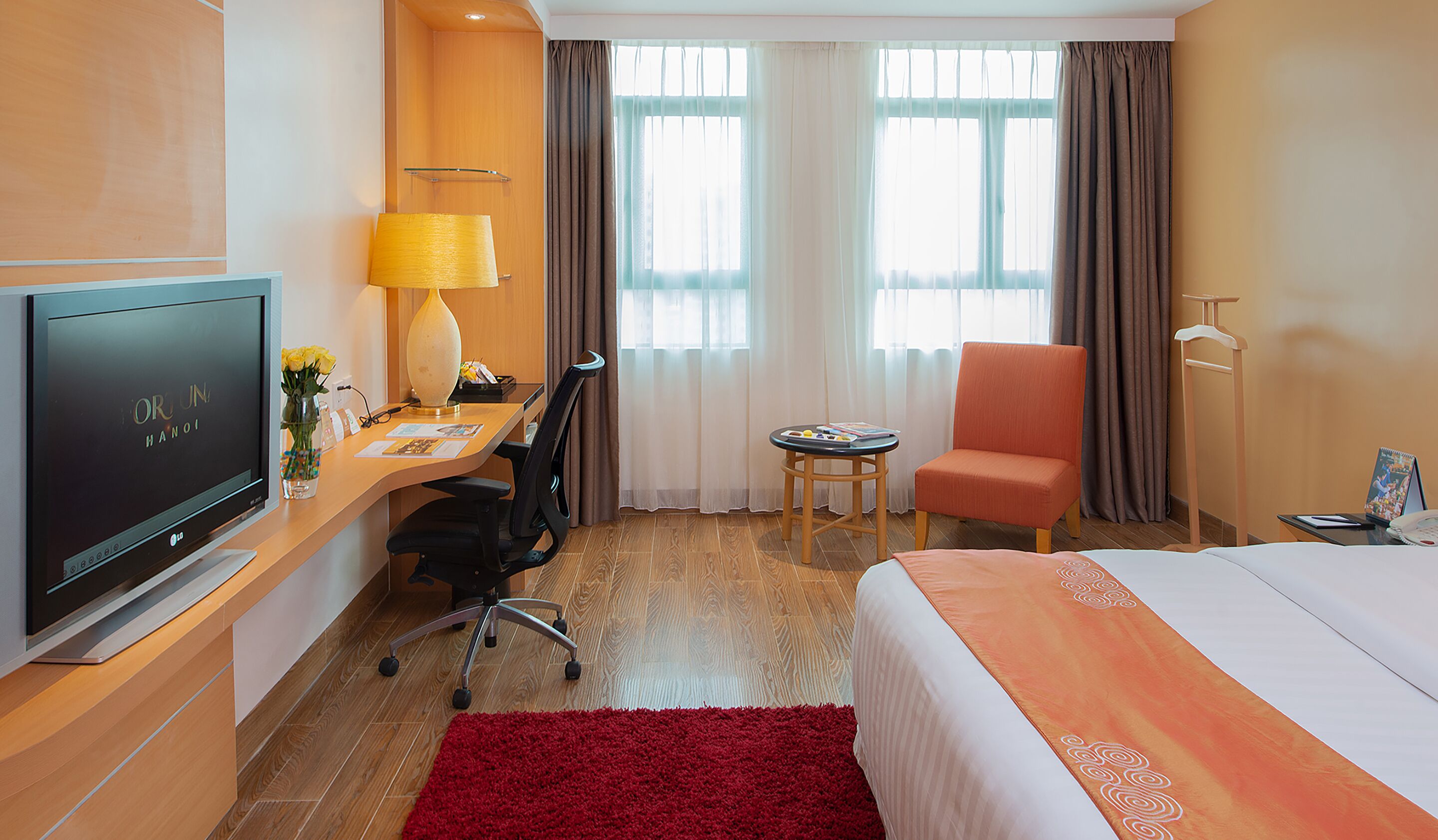 A hotel room with a bed, desk, chair, TV, lamp, and a small table.