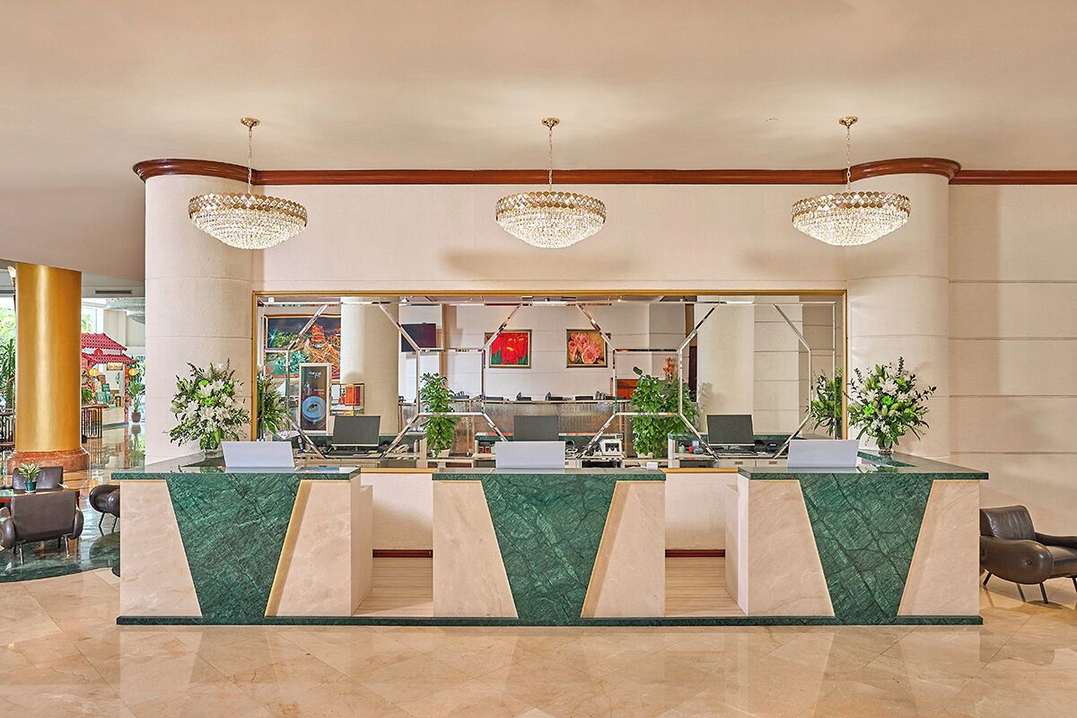 A hotel reception area with a marble counter, three chandeliers, and a large mirror reflecting the interior.