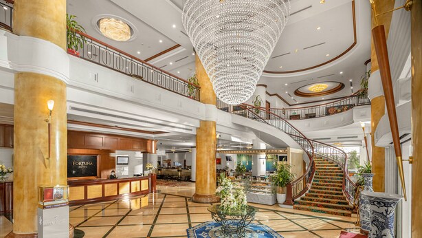 Un amplio vestíbulo de hotel con una imponente lámpara de araña, una recepción y una escalera.