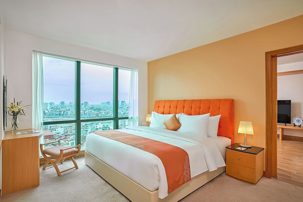A hotel room with a large bed, a desk with a chair, and a view of a cityscape through large windows.