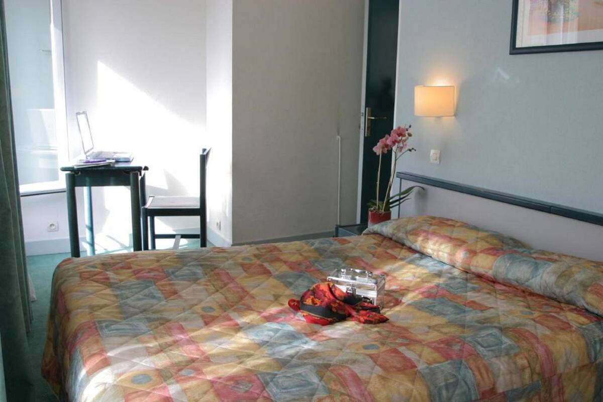A hotel room with a bed, a desk, a chair, a lamp, and a window.